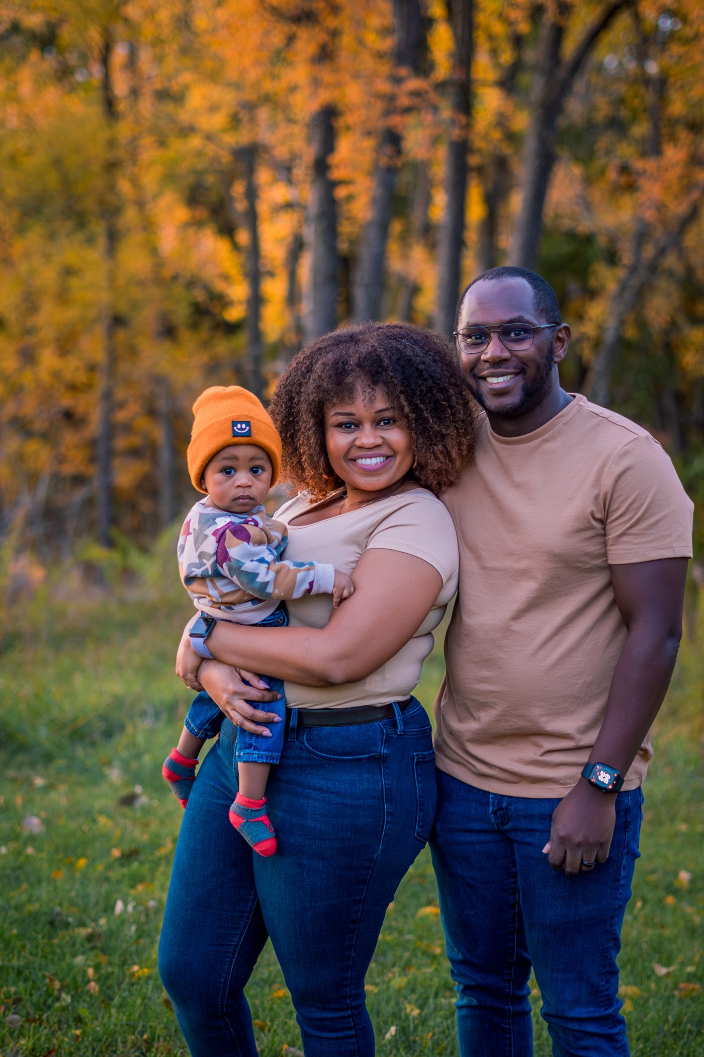 A family of three smiling in front of glowing orange autumn trees